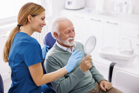 A senior man looks into a mirror to see his smile as a dental assistant looks on, smiling.