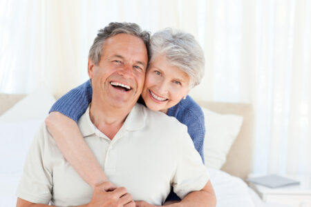 A senior man laughs while a senior woman hugs him from behind, smiling.