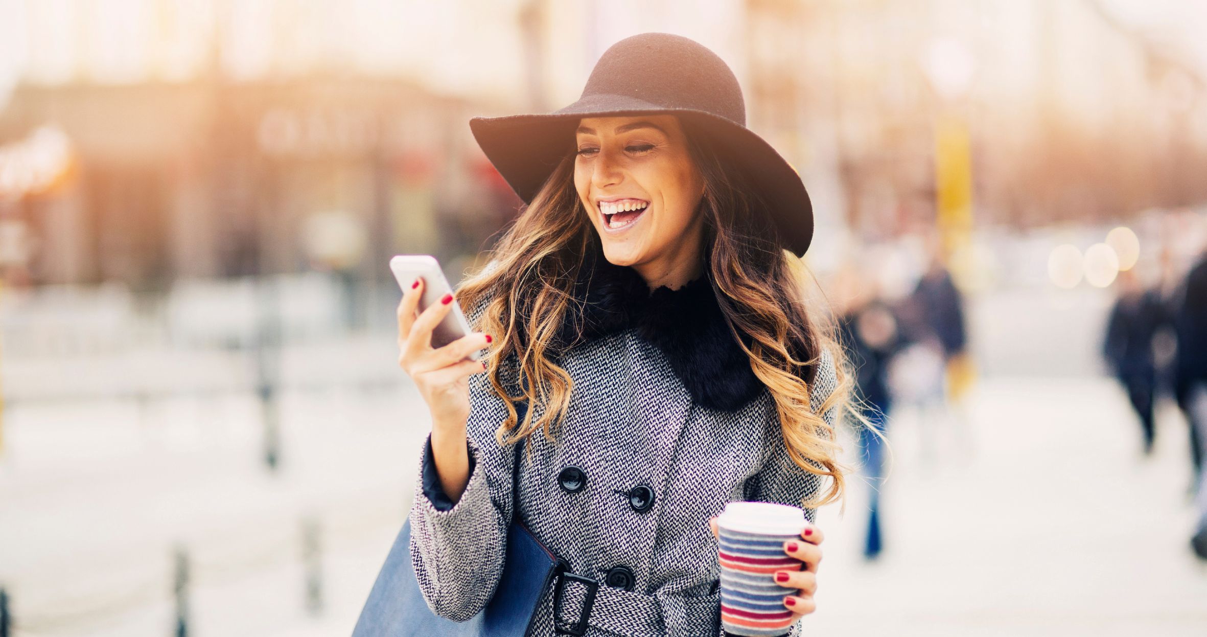 A woman smiles outdoors while looking at her phone and holding a cup of coffee.